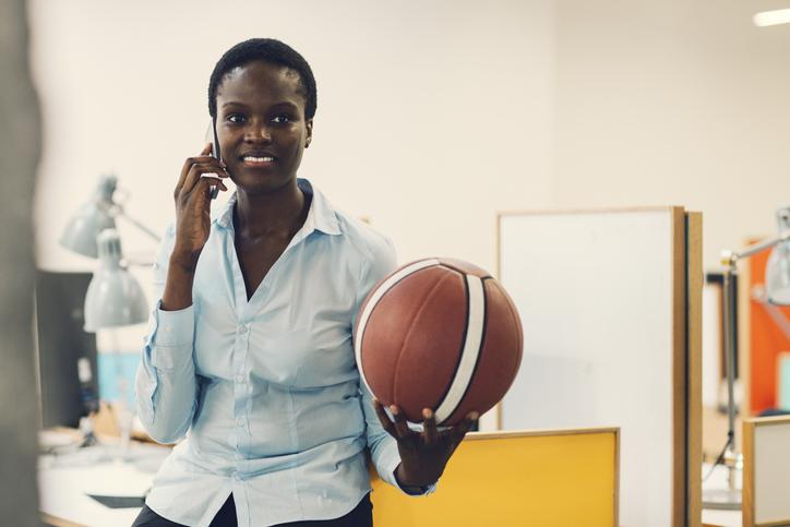 微笑的非洲女商人坐在桌子上，拿着篮球，在现代办公室里打电话. 正面，选择性聚焦在她的笑脸上.